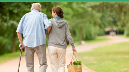 a woman helping and older man with bags as they walk through a park
