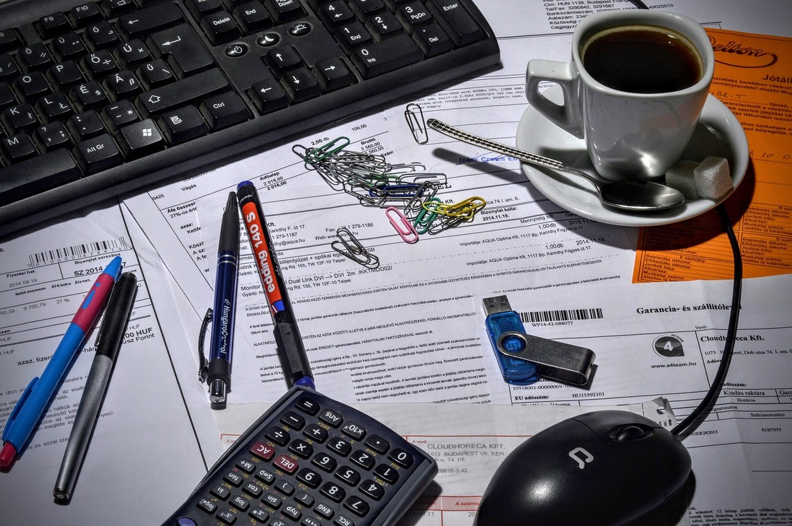 A desk covered in papers with pens, a calculator, mouse, a USB stick and paperclips strewn around with a coffee cup and computer keyboard in the background