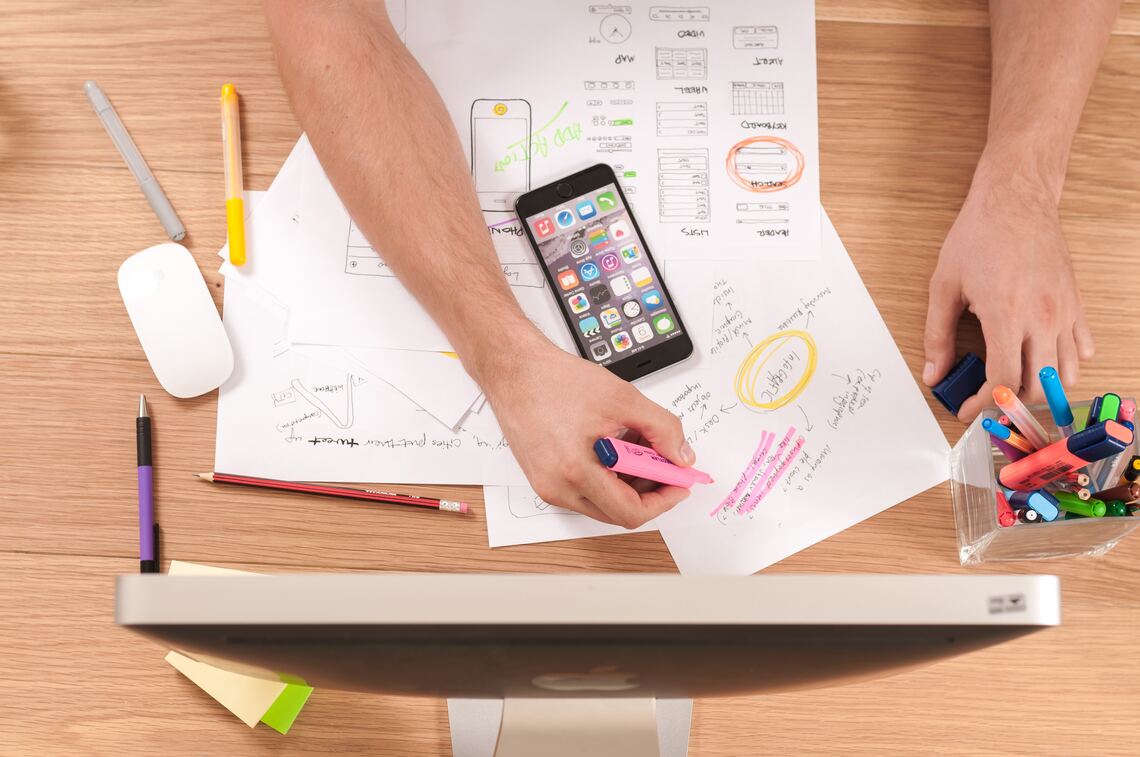 A top down view of a pair of arms in front of a computer highlighting paperwork with a mobile phone, computer, computer mouse and highlighters scattered around the desk