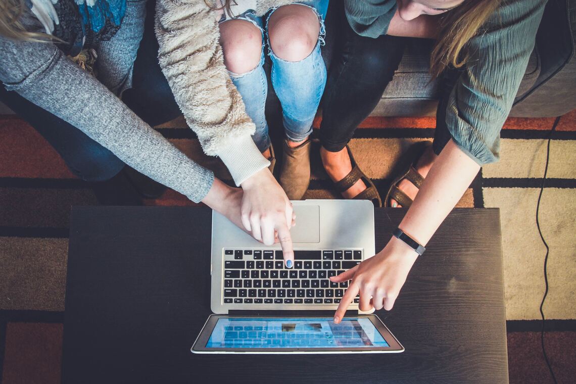 A top down view of 3 people sitting on a sofa and pointing at the screen of a laptop on a table in front of them