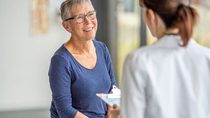 2 women talking, one with her back to the camera wearing a white doctors coat and carrying a clipboard
