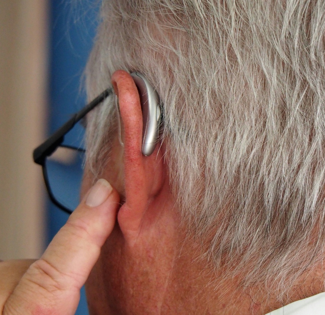 A close up of a side profile of an elderly man's ear with him pointing to a hear aid in his ear with a blue background