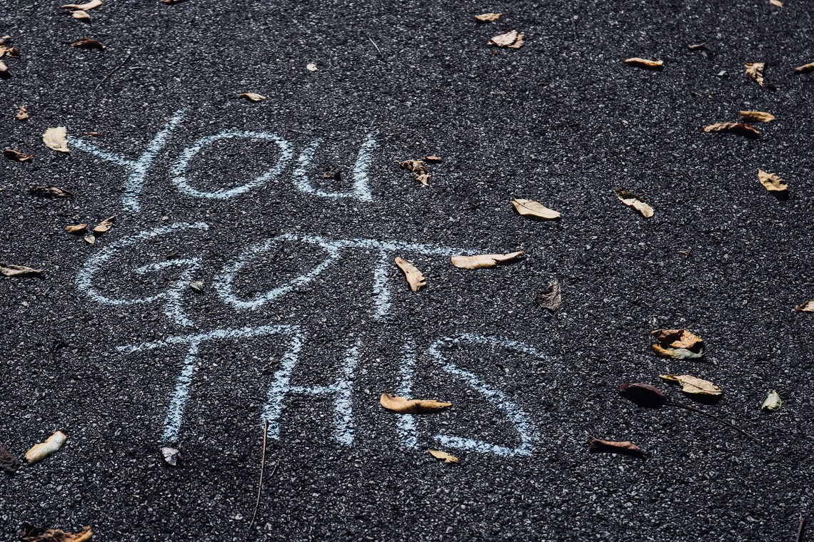 A close up of a road with "you got this" written in white chalk and leaves from trees scattered about