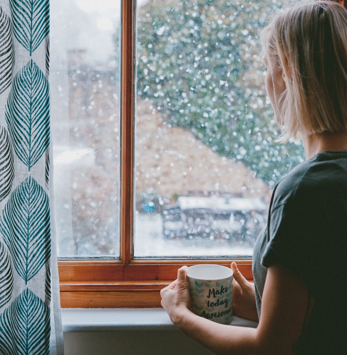 A woman with her back to the camera looking out a window at snow falling with a mug in her hands