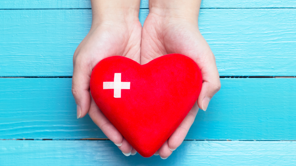 Two hands against a bright blue background holding a red heart shape with a cross in the top left hand corner of the heart
