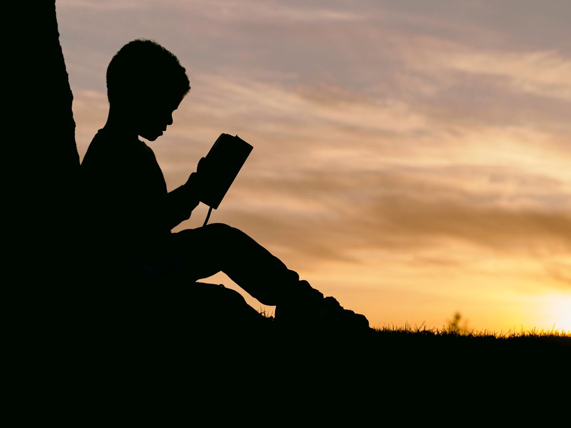 Silohuette of a young child with their back against a tree reading a book outside with the sun setting behind them