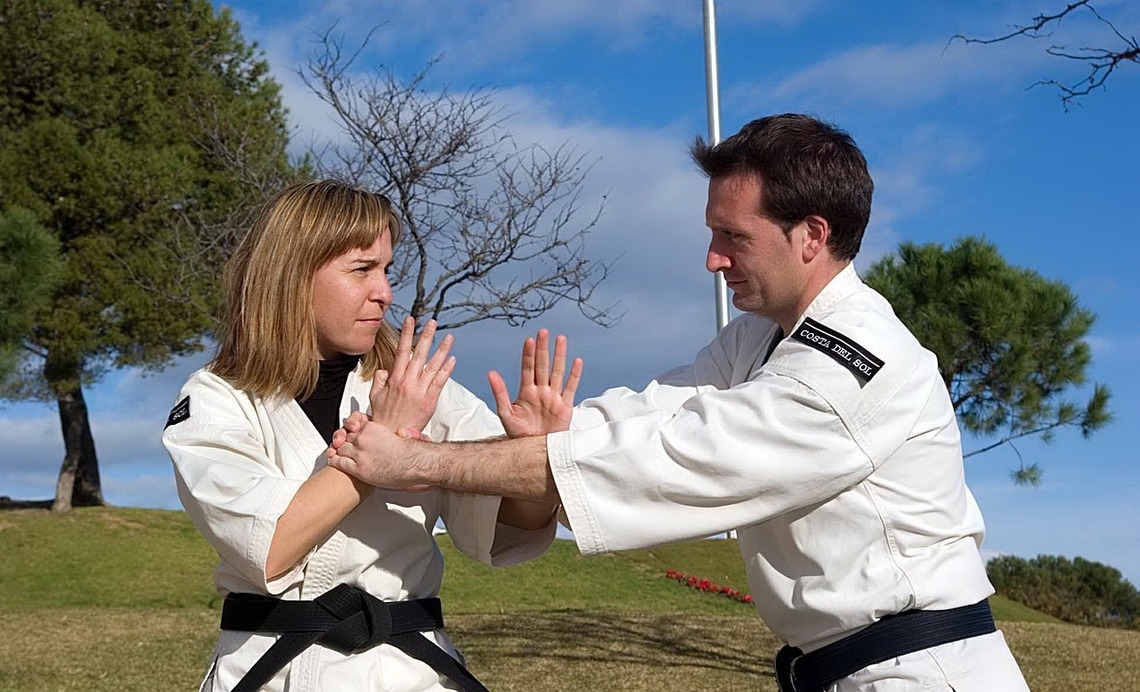 A man holding a woman's wrist in a self-defence stance, both are wearing karate attire, in front of a park setting with green grass, trees, and a blue sky