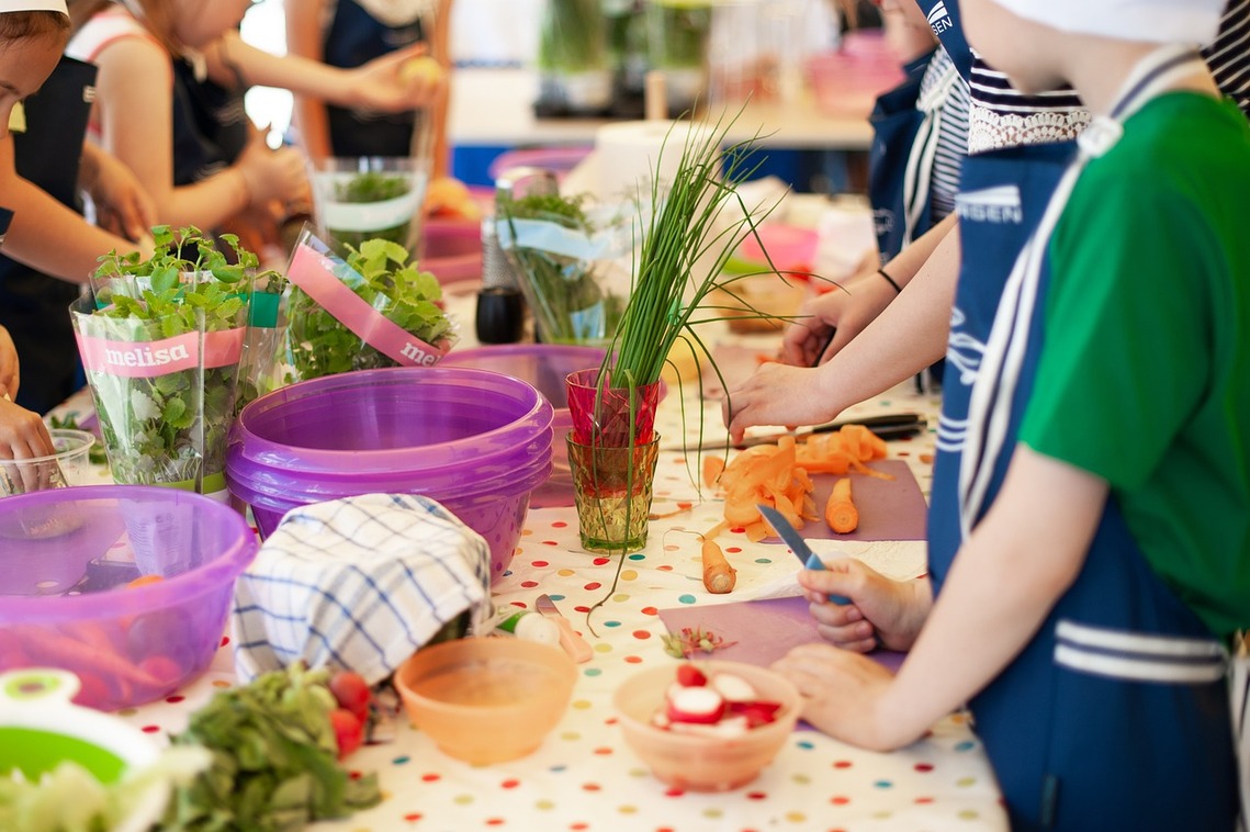 Young children with chef hats and aprons standing along a table with food debris and cooking equipment in front of them
