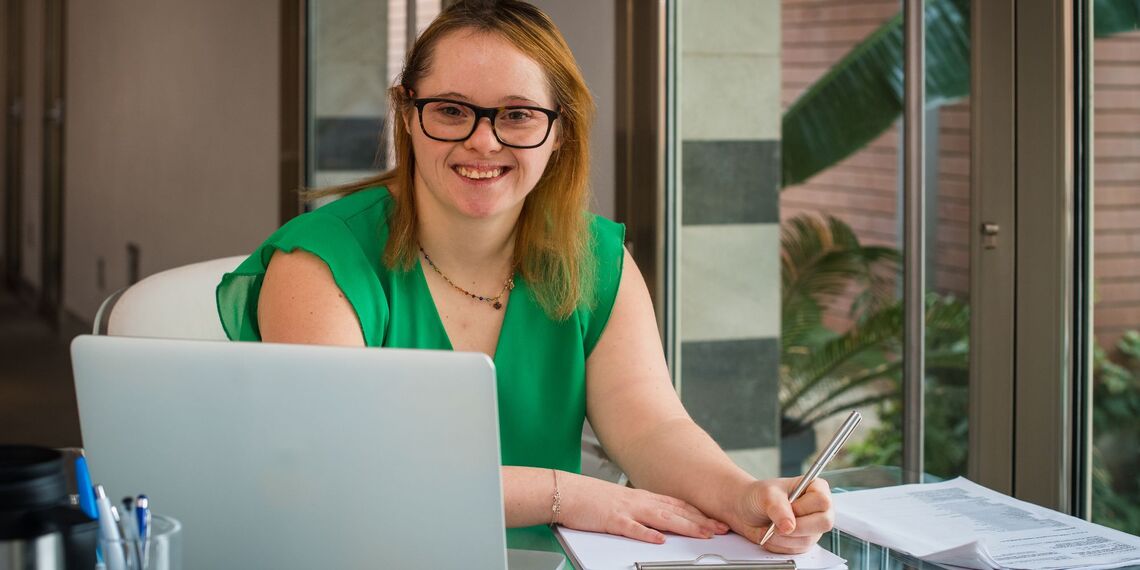 young woman with learning disabilities smiling and working at a computer and paperwork